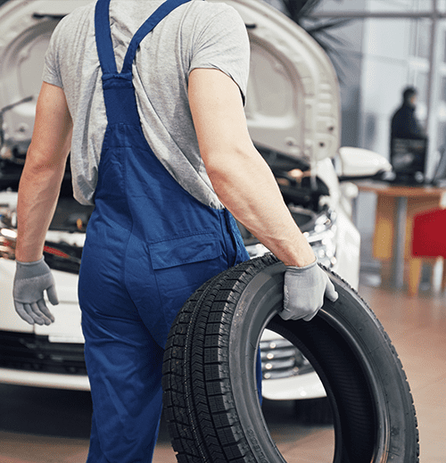 Mechanic carrying a new tyre for installation with car hood open in the background at an auto service shop